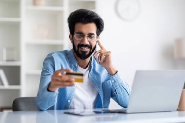 Çevrimiçi sipariş. Kredi kartı tutan ve cep telefonuyla konuşan mutlu Hintli bir adam ofis masasında otururken, doğu erkeği müşteri destek müdürüyle sohbet ederken, alışveriş yaparken, kopyalama alanı yaparken,
