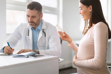 Hamile bir kadının muayenesi sırasında sağlık raporlarına yazı yazan bir erkek doktor elinde bir kavanoz hap tutan bir kadın, seçici odaklanma. Kadın doğum uzmanı, çocuk bekleyen bir kadına danışmanlık yapıyor.
