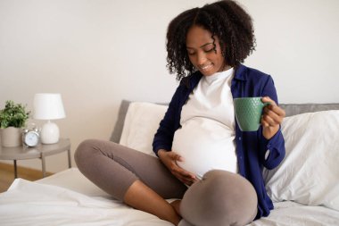 Bin yıllık mutlu Afro-Amerikan hamile kadın, koca göbekli, bir fincan çay içen, yatak odasının içinde bebeğin hareket ettiğini hisseden. Bekleyen çocuk, annelik ve aile, evde dinlenme
