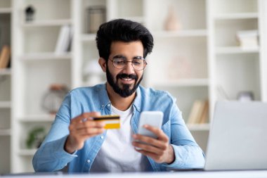 Kolay ödemeler. Evde akıllı telefon ve kredi kartı kullanan gülümseyen Hintli adam, masasında oturan mutlu Doğu 'lu genç adam, uzaktan çekimde çalışırken internet üzerinden cep telefonu alışverişi yapıyor,