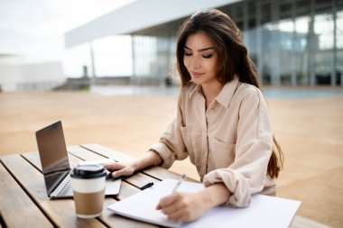 Çoklu görev, eğitim ve çalışma. Üniversitede bilgisayarlı gülümseyen Asyalı genç bayan öğrenci kahve fincanı ile masada, açık havada. Uzaktan ders, modern eğitim, seminer ve sınav hazırlığı için aygıt