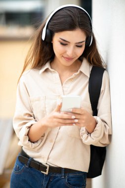 Sırt çantalı, kulaklıklı gülümseyen Asyalı genç kız öğrenci, akıllı telefonla konuşuyor, açık havada, dikey. Öğrenim uygulaması, öğrenme aygıtı, ses dersleri ve müzik, modern eğitimden ayrıl