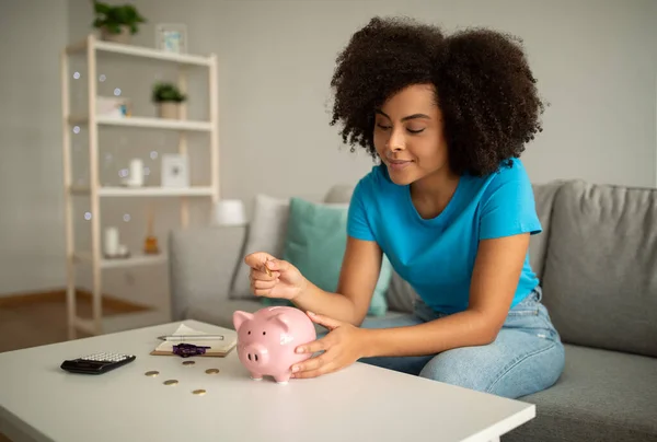 Smiling young black curly woman puts money in piggy bank, counts profit in living room interior. Finances for purchase of dream, study, savings at home, accounting and economic