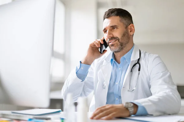 快乐的专业男医生在手机上交谈 与病人进行流动对话 在网上提供建议 坐在诊所的工作场所 — 图库照片