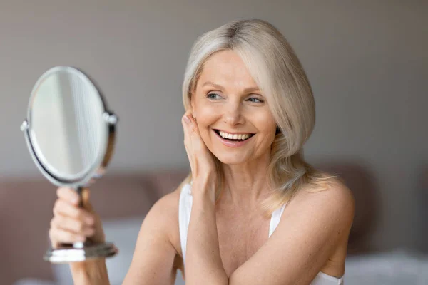 老化的自然美概念 快乐的老年女性看着镜子 触摸着自己的脸 微笑着反思 享受着新的护肤产品 — 图库照片