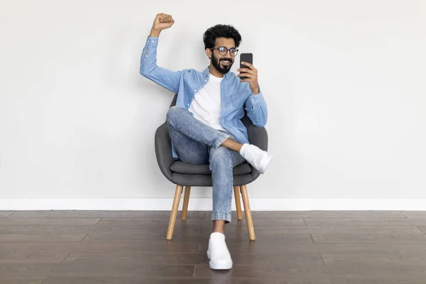 Vreugdevolle Jonge Indiase Man Zit Stoel Met Smartphone Vieren Van — Stockfoto