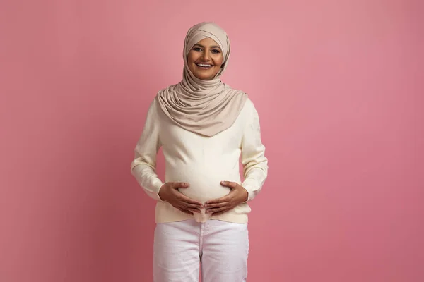 ピンク色のスタジオを背景にポーズをとりながらベリーを抱くムスリム妊婦さん カメラで赤ちゃんのお尻を愛撫しながら微笑む幸せなアラビア語の妊婦さん コピースペース — ストック写真