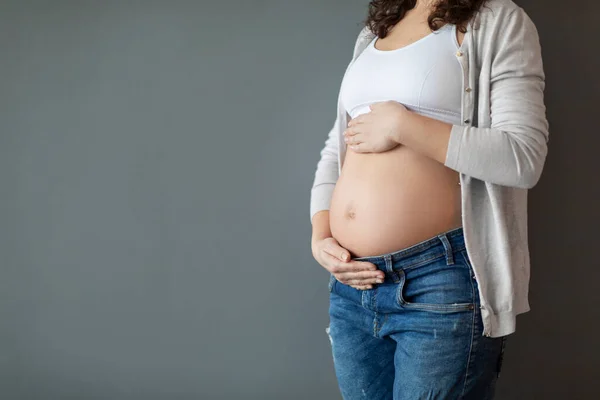 出産の概念 灰色のスタジオの背景に立っている間に手で腹を抱えている若い妊娠中の女性の作物のショット 認識できない妊婦は赤ちゃんの塊を抱きます スペースをコピー — ストック写真