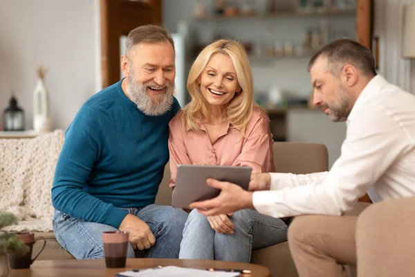 Professional Agent Showing Tablet Computer To Excited Senior Couple Offering Digital Services Sitting On Couch At Home. Real Estate Buyers Choosing House Property With Specialist. Selective Focus