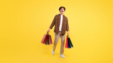 Başarılı bir alışveriş. Renkli kâğıt torbaları taşıyan mutlu genç adam, Satış sezonunun tadını çıkartıyor Sarı Stüdyo Arkaplanı Üzerine Poz Veriyor. Ticaret ve Perakende. Panorama, Tam Uzunluk Görüntüsü