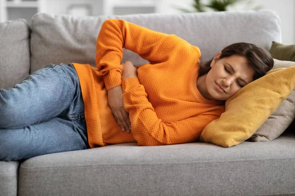 患有胃痛 躺在沙发上 抱着肚子 忍受肚子痛和不适 月经抽筋 复制空间的年轻印度妇女 — 图库照片