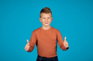 Yakışıklı, gülümseyen ergen çocuk kameraya başparmak hareketi yapıyor, olumlu ergen erkek çocuk onay jesti gösteriyor, mavi arka planda dururken iyi bir teklif sunuyor, boşluğu kopyalıyor.