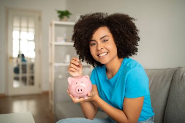 Gülümseyen milenyum kıvırcık saçlı zenci kadın kumbaraya bozuk para koyar. Oturma odasının içinde rüya görmek için. Para finansmanı, kâr, evde muhasebe, ekonomi, tasarruf ve satın alma kredisi