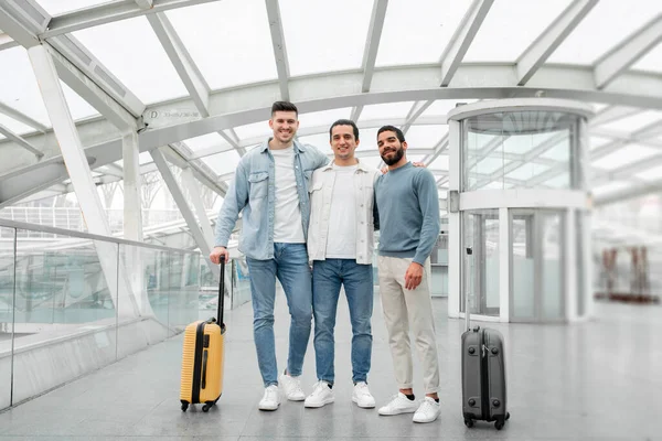 stock image World Wanderers. Three Friends Men With Travel Suitcases Embracing Posing In Airport, Exploring Globe Together Traveling On Vacation Abroad. Modern Tourism And Transportation. Full Length Shot
