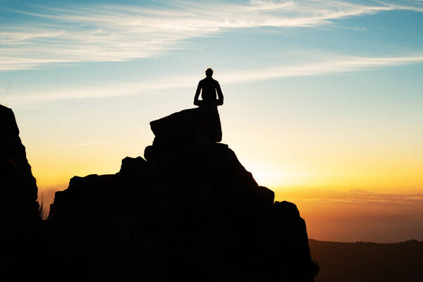 Silhouette Of Man Sitting In Lotus Position On Top Of Mountain Cliff Meditating Looking At Sunset Outdoor. Rear View Of Male Hiker Tourist Resting After Hike. Tourism Concept