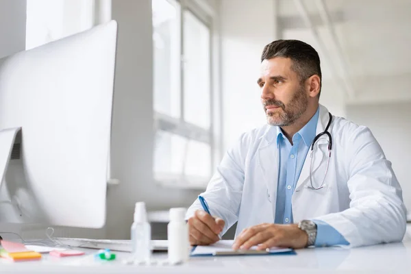 Electronic health system. Male doctor working on computer and writing prescriptions while sitting at desk in office. Physician man wearing medical coat, consulting patients online in clinic
