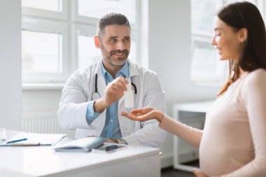 Hamilelik sağlığı. Genç hamile bir kadın beslenme uzmanını ziyaret ediyor, elinde ilaç şişesi olan bir erkek doktor klinikteki kadın hastaya danışmanlık yapıyor, vitamin veya takviye ilaç veriyor.
