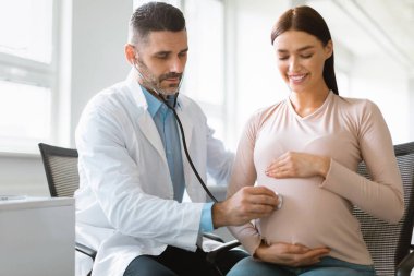 Hamile kadın ve bebeğin sağlığı. Hastanedeki genç bayan randevusu sırasında bebek kalbini dinleyen bir erkek doktor. Genç bayan hasta jinekoloğunu klinikte ziyaret ediyor.