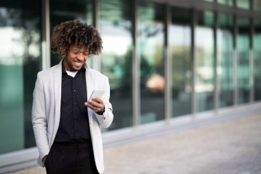 Ofis binasının dışında yürüyen, akıllı telefon kullanan, mesaj yazan, gülümseyen, internette harika haberler okuyan, boşluğu kopyalayan mutlu siyahi işadamı.