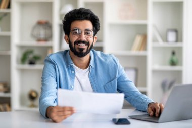 İçişleri Bakanlığı 'nda çalışan yakışıklı Hintli serbest çalışan, laptopla masada oturan ve mali belgeleri kontrol eden, kameraya bakan, boş alana bakan, gülümseyen genç bir doğulu.