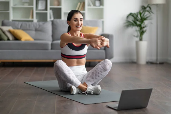快乐而又活泼的漂亮的黑发女子坐在瑜伽垫上 看着笔记本电脑屏幕 手拉手在家里 上在线健身课 抄袭空间 — 图库照片