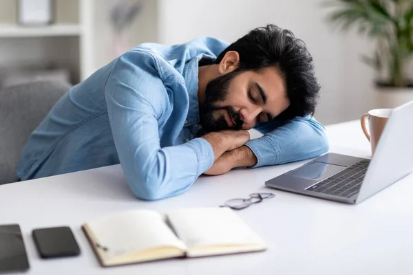 Overworking Concept. Tired Young Indian Man Napping At Desk With Laptop, Eastern Male Freelancer Exhausted After Working On Computer At Home Office, Placing Head On Hands, Closeup Shot
