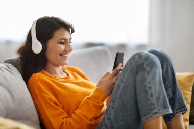 Ev eğlencesi. Evdeki akıllı telefon ve kablosuz kulaklık kullanarak gündelik koltukta oturan neşeli, güzel Hintli kadın video içeriği veya filmi internetten izliyor.
