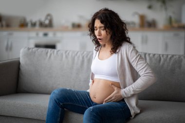 Braxton Hicks Contractions. Young Pregnant Woman Having Abdominal Pain At Home, Stressed Expectant Mother Sitting On Couch In Living Room And Touching Belly, Feeling Unwell, Copy Space clipart