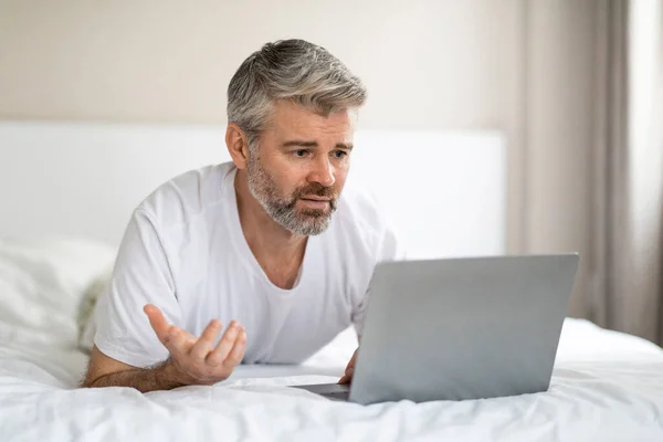 中年白发苍苍的中年男人在家里躺在床上休息的时候 一边用笔记本电脑 一边看着现代电脑屏幕 一边摆弄姿势 一边看垃圾邮件 或者软件有问题 一边抄袭空间 — 图库照片