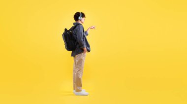 Sırt çantalı, kulaklık takan, müzik dinleyen ve görünmez düğmeye basan ya da sarı stüdyo arka planında boş yer gösteren genç adam görüntüsü. Panorama