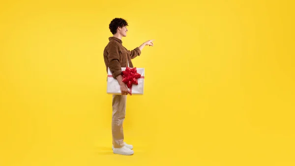 礼物的乐趣 年轻人拿着一个包装好的礼品盒 一边指指点点着手指 一边假装敲响着无形的门铃 把黄色的画室背景挂在那里 礼品派递服务 — 图库照片