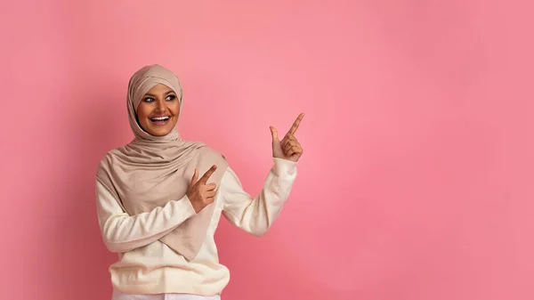 特价优惠喜气洋洋的穆斯林女性带着两只手指在复印空间边指指点点 兴奋快乐的伊斯兰女性在粉红工作室的背景下展示广告的自由位置 — 图库照片