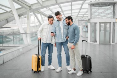 Bağlantılı Gezginler. Akıllı telefonlar kullanan, bavullarıyla ayakta duran, seyahat başvurusu yapan ve havaalanı terminalinde uçuş rezervasyonu yapan üç turist grubu. Tam Uzunluk