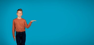 Bunu dene. Gülümseyen Kafkasyalı Çocuk Boş Avucunda Görünmez Nesne Gösteriyor, Sevimli Erkek Çocuk Reklam İçin Bedava Yer Gösteriyor Ya da Mavi Arkaplan Üzerinde Teklif Ediyor, Panorama