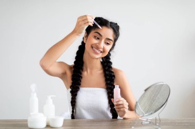 Cilt bakım konsepti. Güzel Hintli kadın, evde aynanın önünde otururken Düşükle yüz masajı yapıyor, Gülümseyen Doğulu kadın cildini nemlendiriyor, kişisel bakım rutininden zevk alıyor.