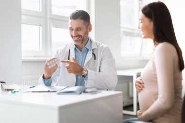 Doğum öncesi bakım ve tedavi. Mutlu erkek doktor, hamile bayan için vitamin reçetesi yazıyor. Elinde ilaçlarla barutu tutuyor ve anne adayına tablet veriyor.