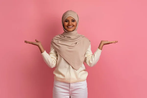 做出选择 穿着头巾 面带微笑的穆斯林女士 用她那空空的手掌做天平 快乐的年轻的伊斯兰女人 一边站在粉红的工作室背景上 一边比较各种选择或做广告 一边抄袭空间 — 图库照片