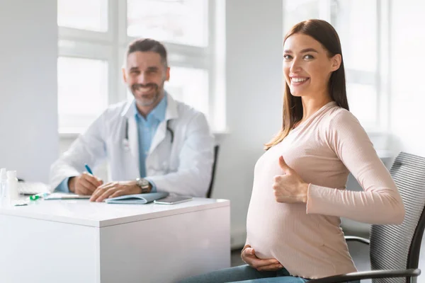 怀孕情况检查 满意的年轻孕妇在镜头前微笑 竖起大拇指 坐在医生咨询处 推荐高质量的妇女保健医疗服务 — 图库照片