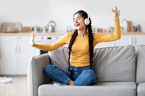 自宅で楽しい 無線電話で彼女のお気に入りの音楽を聞いている陽気な若いインドの女性の肖像 リビングルームでソファに座っている間に手で踊るケアフリー東の女性 — ストック写真