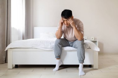 Mali stres: Üzgün, orta yaşlı, evde pijama giyen, kafası dokunan, maddi durumla, borçla, işsizlikle, beklenmedik harcamalarla boğuşan Asyalı adam.