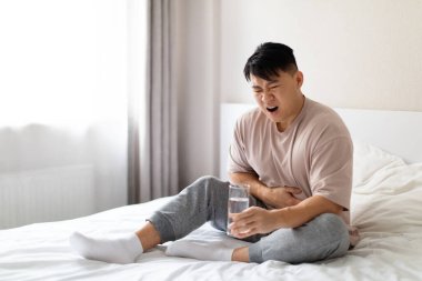 Mutsuz, orta yaşlı Çinli bir adam yatakta oturuyor ve karnına dokunuyor, sabahları mide ağrısı çekiyor, pijama giyiyor, elinde bir bardak su tutuyor. Gıda zehirlenmesi, ishal konsepti