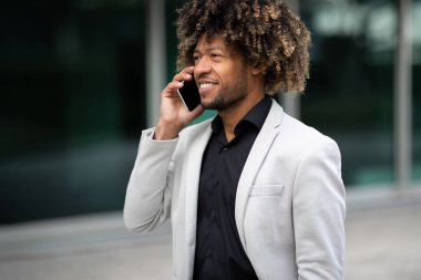 Neşeli Afro-Amerikan orta yaşlı iş adamı akıllı telefondan konuşuyor, modern ofis merkezinin yakınında dışarıda yürüyor. Siyah erkek girişimci telefonda konuşuyor.