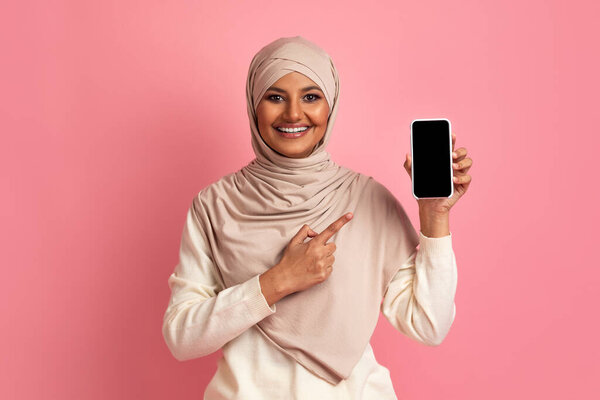 Smiling Muslim Lady Pointing At Blank Smartphone With Black Screen In Her Hand, Islamic Woman In Hijab Recommending New Mobile Application While Standing On Pink Studio Background, Mockup