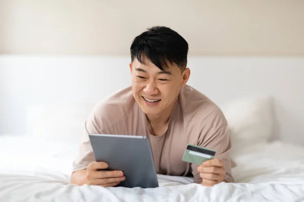 电子商务 在线零售 快乐的中年男子 英俊的亚裔男子 喜欢从家里上网购物 躺在床上 使用现代Pc数码平板电脑和银行卡 穿着睡衣 复制空间 — 图库照片