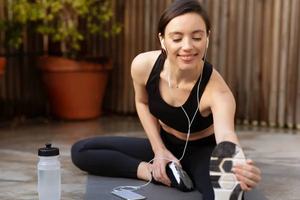 Glad young caucasian slender woman in sportswear and headphones doing stretching for legs on mat with bottle of water outdoor. Music for sports and warmup, workout, health care in city
