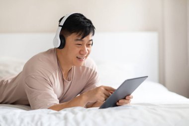 Heyecanlı, neşeli, orta yaşlı, pijamalı Çinli adam evde yatarken, kablosuz stereo kulaklık ve dijital tablet kullanıyor, müzik dinliyor, video içeriği izliyor, yer kopyalıyor.