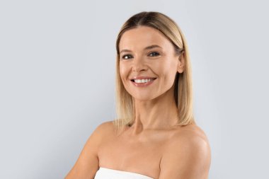 Portrait of cheerful attractive half-naked middle aged blonde woman smiling at camera, posing on grey studio background, demonstrating glowing perfect skin, copy space. Cosmetology concept