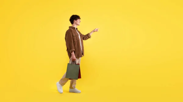买东西的广告 微笑的年轻人背着纸袋 在空旷的空间为文字广告指出手指 站在黄色工作室的背景上 — 图库照片