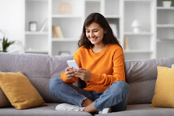 快乐迷人的长发年轻女性 身穿休闲装 坐在沙发上 双腿翘起 使用手机和微笑 查看最新的移动应用程序 复制空间 — 图库照片