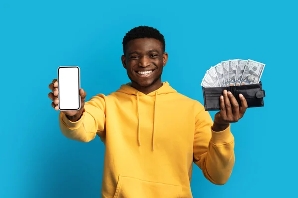 在线贷款 快乐英俊的年轻黑人 穿着黄色的帽衫 拿着白色的空白屏幕 钱包里装满了钞票 蓝色的工作室背景 模仿着现代手机 — 图库照片